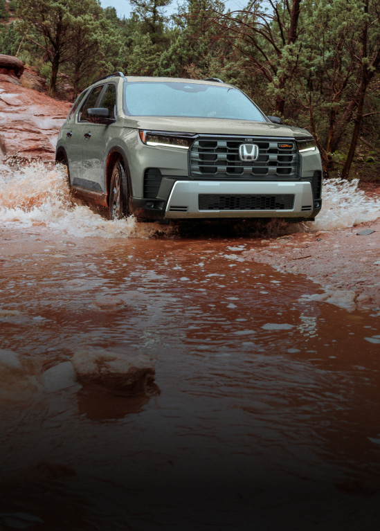 2026 Honda Pilot TrailSport in Ash Green Metallic front driving through a rocky stream in a forest (Full-Size SUV).